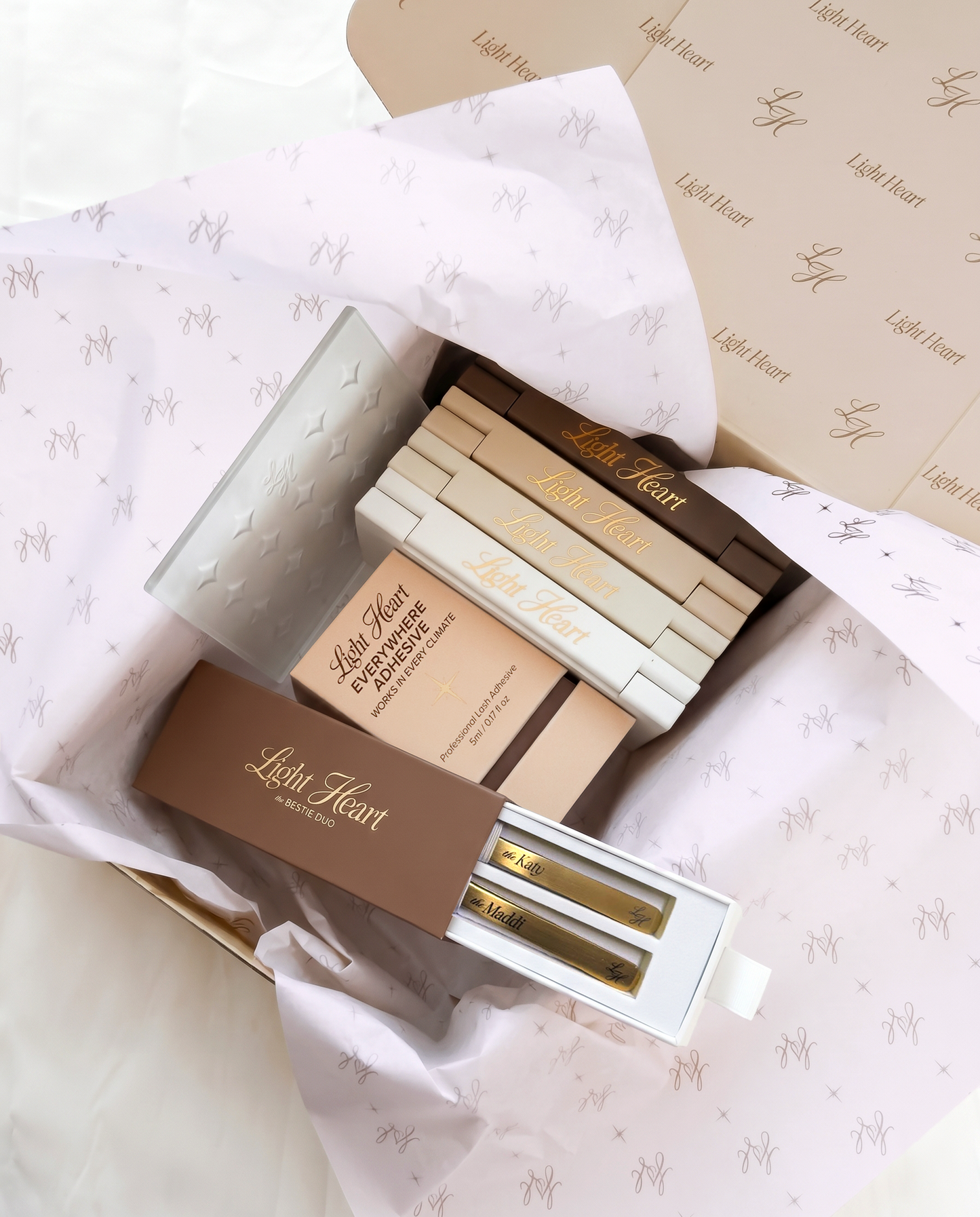 Light Heart Lashes unboxing flat lay showing the Bestie Duo tweezer set with Katy and Maddi tweezers, stacked lash trays, Everywhere Adhesive, and branded LH tissue paper inside an open Light Heart shipping box