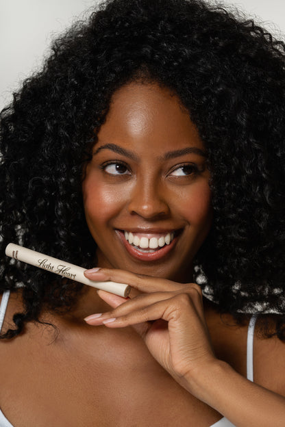 Smiling woman with natural curly hair holding Light Heart Brow Growth Serum, showcasing a nourishing eyebrow treatment for fuller, healthier-looking brows.
