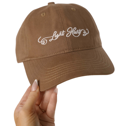 Light Heart Lashes brown dad hat with white embroidered script logo, held up by hand